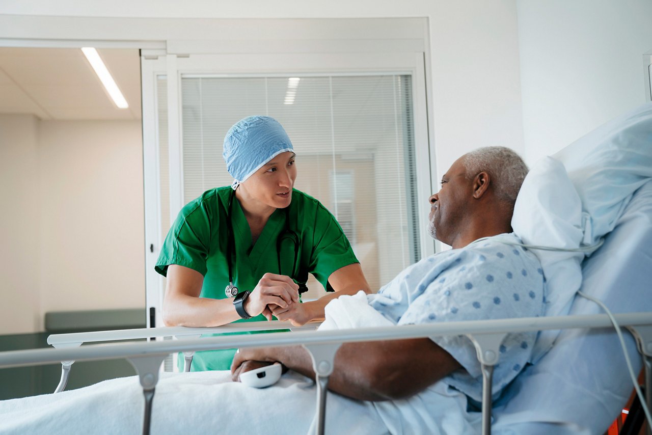A male clinician in scrubs and a surgery cap talking to an older male patient who is smiling while lying in a hospital bed