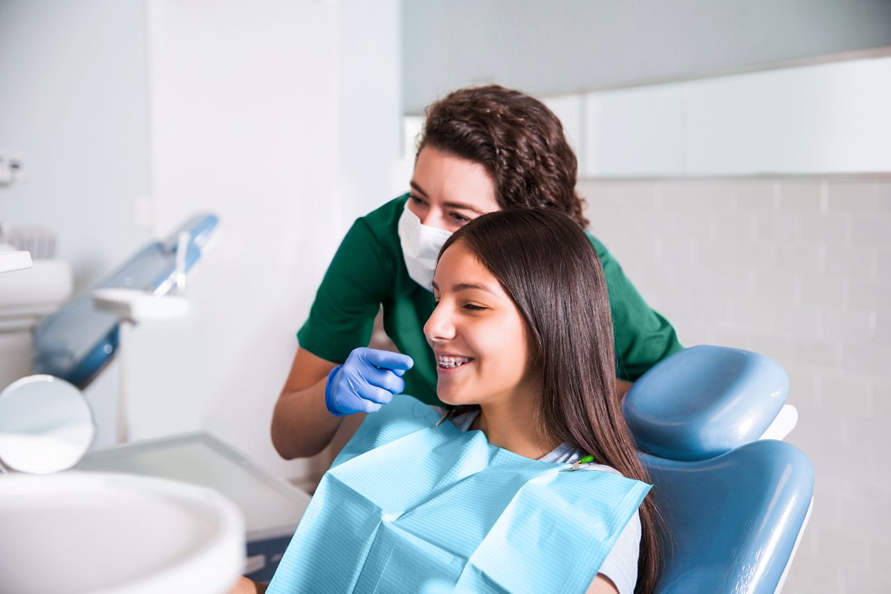 In scrubs by a tray table, a seated orthodontist smiles at the camera.