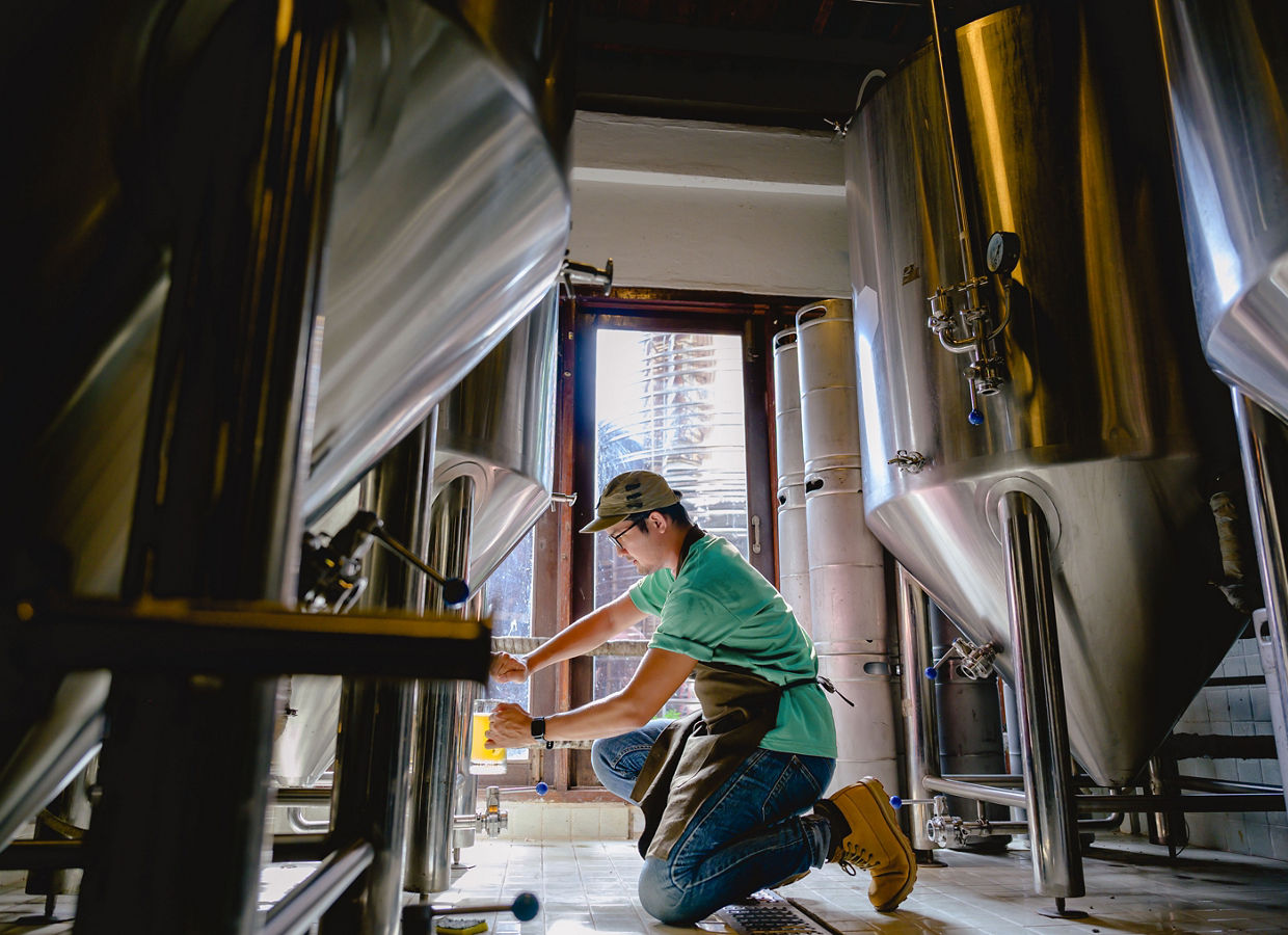 Young male brewer kneels on one knee to dispense beer into a glass to check the taste and color in a craft brewery.