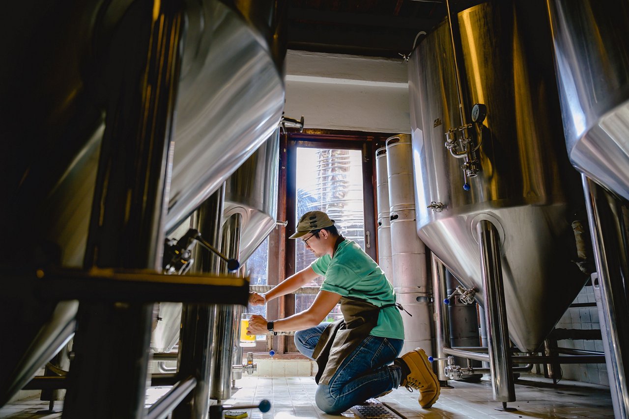 Young male brewer holding a glass of beer in hand under the spout of a brew kettle in a production facility.