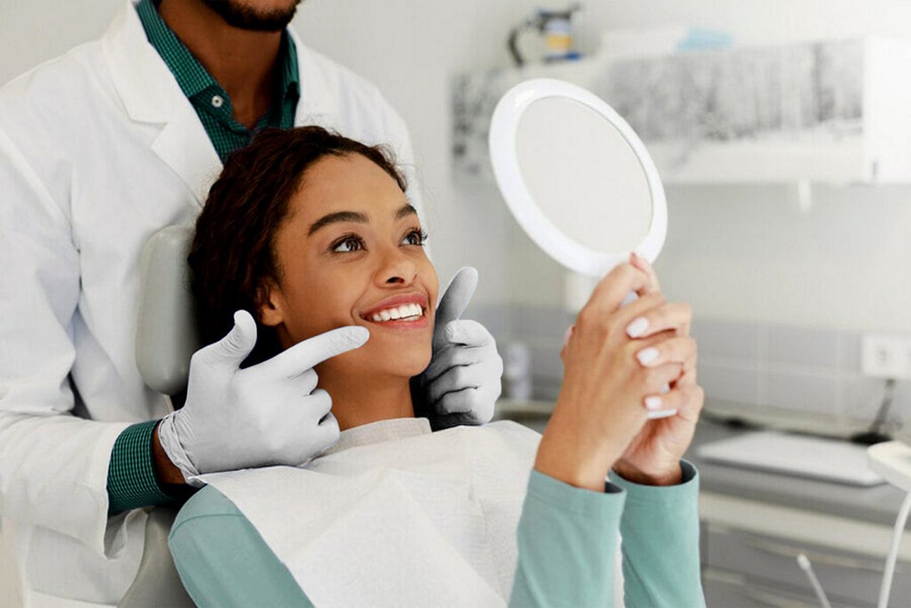 An image of a women looking into a hand mirror at her teeth while her orthodontist is pointing towards her teeth