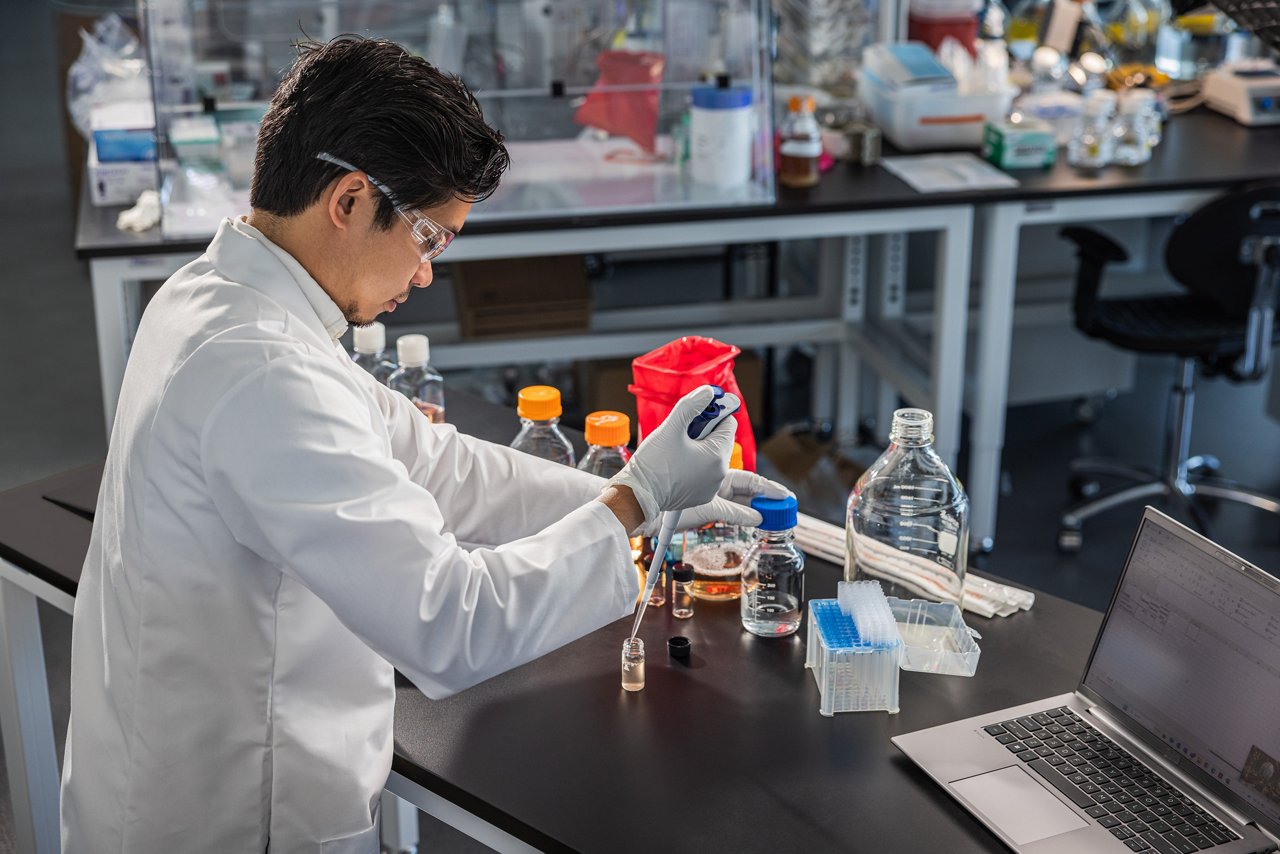 Image of a laboratory worker testing liquid on a bench with a laptop for data entry.