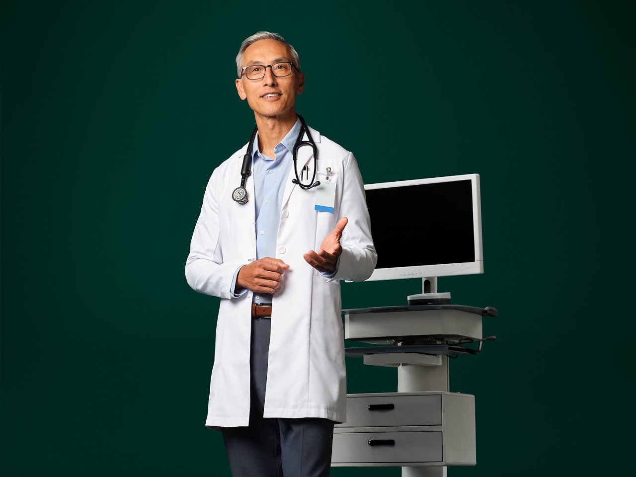 Un médecin avec un stéthoscope autour du cou debout à côté d’un appareil ambiant comme s’il parlait à un patient.