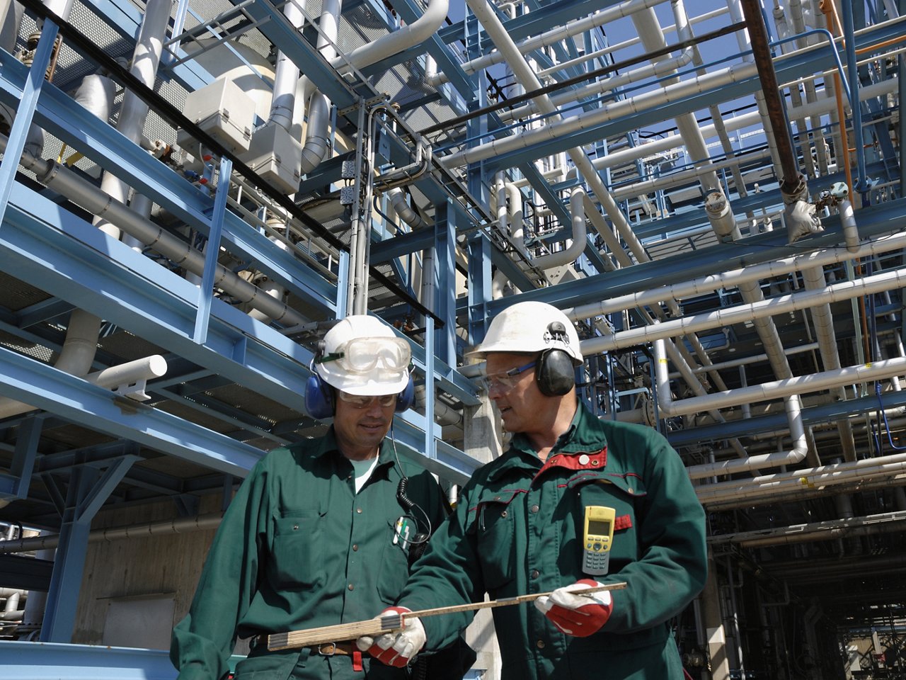 two engineers with hard hats standing in front of a refinery and miles of pipelines