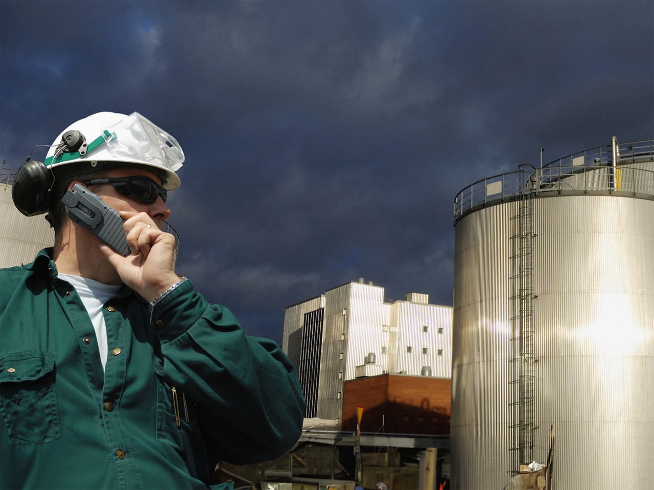 oil and fuel storage tanks with worker in foreground wearing hardhat and using cellphone