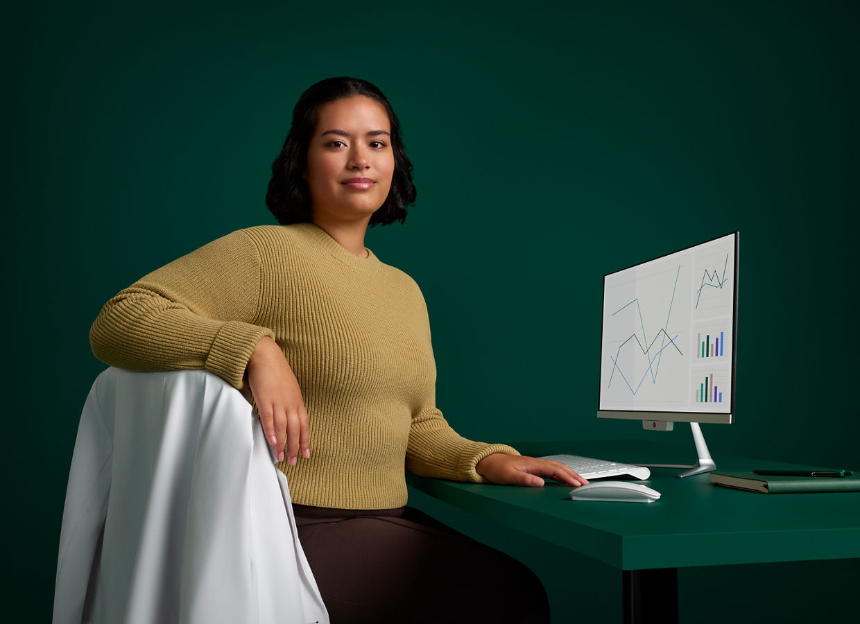 Portrait of an infection prevention specialist sitting at her desk with computer on a dark green background.