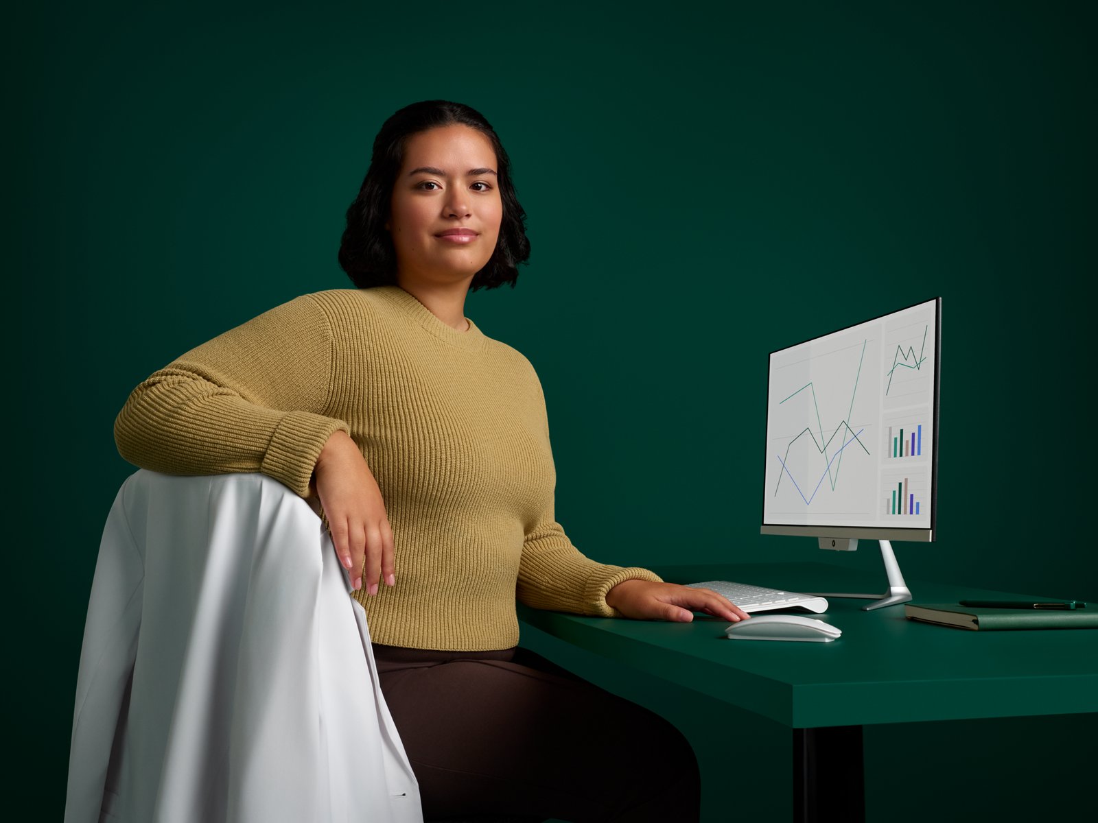 Portrait of an infection prevention specialist sitting at her desk with computer on a dark green background.