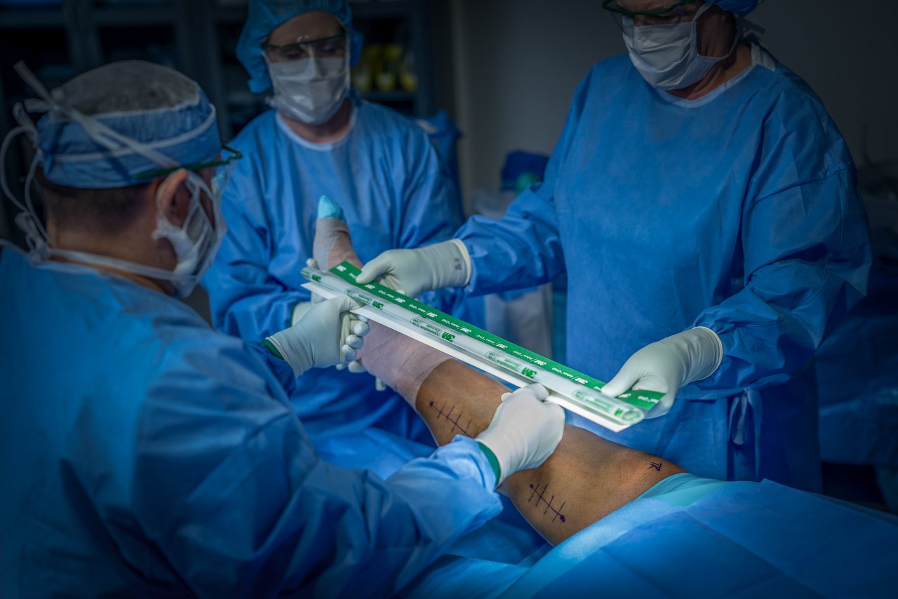 Two OR nurses hold onto the edges of the closed Ioban™ CHG Incise Drape over the patient's leg while another OR nurse holds the leg up.