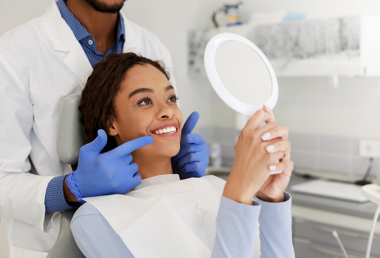 A woman patient in the chair smiles into a hand mirror as the dentist points at her white teeth.