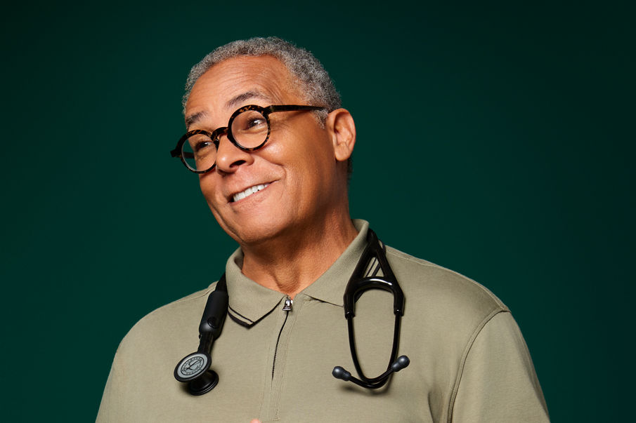 Portrait of a male primary care doctor with Littmann stethoscope around his neck on a dark green background.