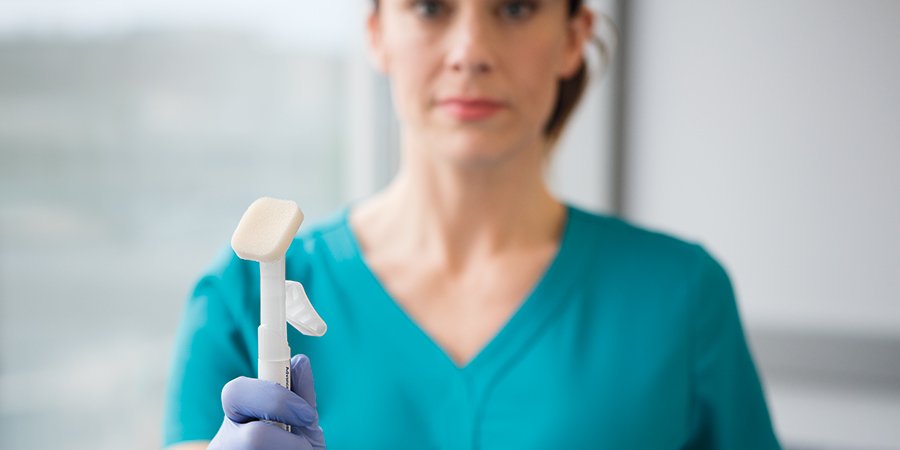 Odak noktasında Cavilon Advanced uygulayıcı ve uygulayıcıyı eldivenli eliyle tutan hemşirenin bulanık olduğu stilize çekim. Cavilon Kahraman Görseli, Kırpma 900x450, rgb, jpg