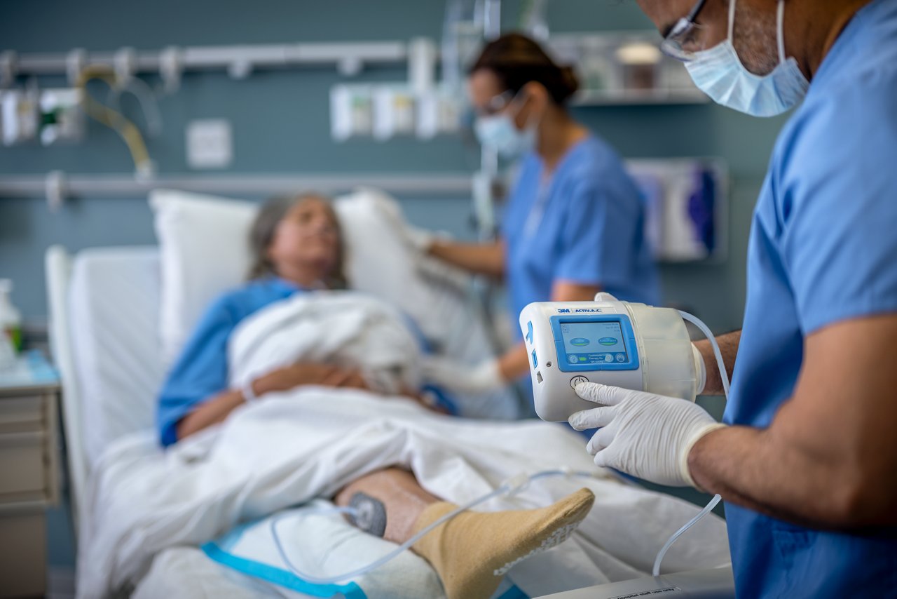 Patient in ICU with a venous leg ulcer with 3M™ Dermatac™ Drape and 3M™ V.A.C.® Granufoam™ Dressing  receiving 3M™ ActiV.A.C.™ Therapy. Clinicians with white gloves and blue gowns monitoring the patient., rgb, tif
