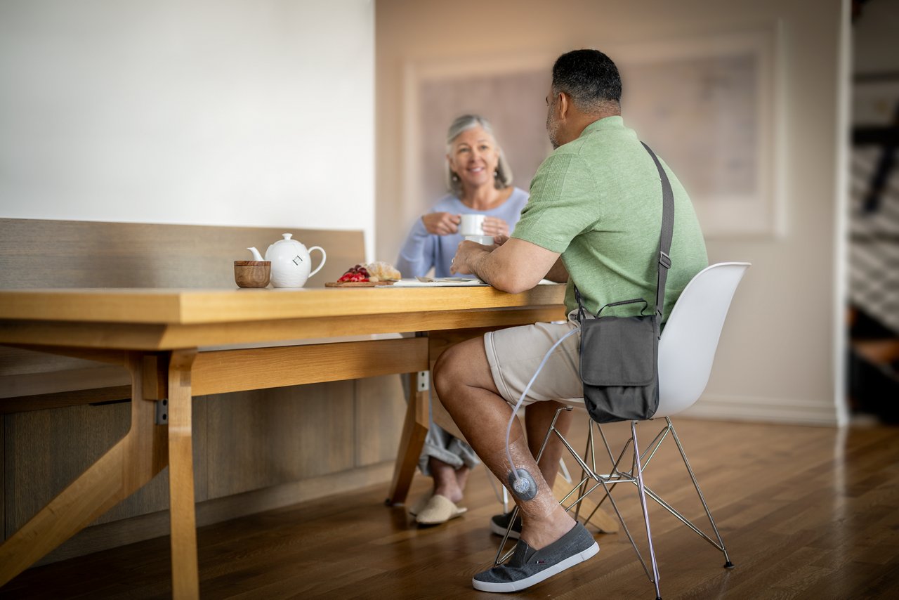 Patient at home sitting at table with friend wearing 3M™ V.A.C.® Peel and Place Dressing and 3M™ ActiV.A.C.™ Therapy Unit in pouch. Includes 3M™ SensaT.R.A.C.™  Pad.