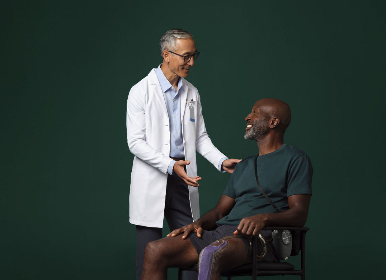Male surgeon in white coat standing next to and talking with a seated male patient who has a Prevena therapy device on his leg