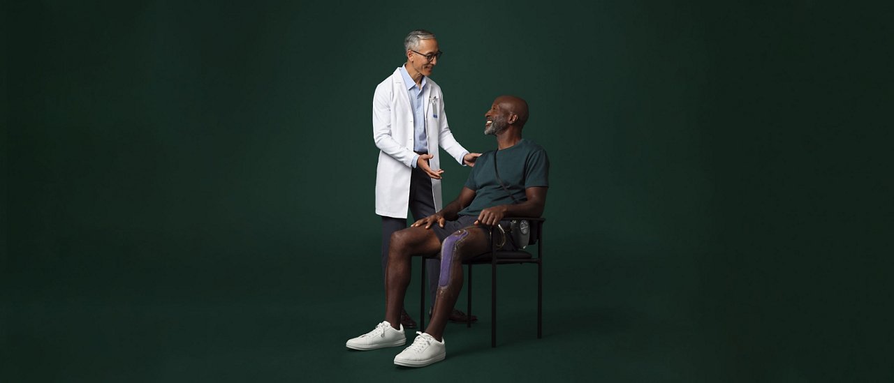 Mandlig kirurg i hvid kittel, der står ved siden af og taler med en siddende mandlig patient, som har en Prevena-behandlingsenhed på sit ben