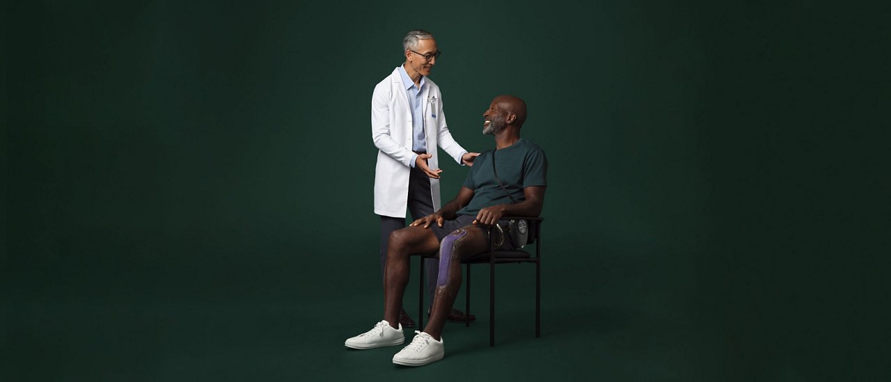 Male surgeon in white coat standing next to and talking with a seated male patient who has a Prevena therapy device on his leg