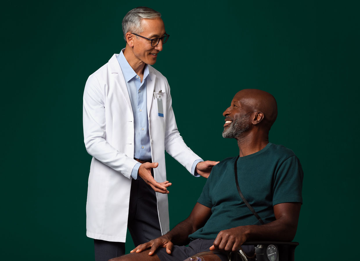 Um cirurgião de jaleco branco em pé ao lado de um paciente sentado que está conversando e que possui um dispositivo de terapia Prevena na perna.