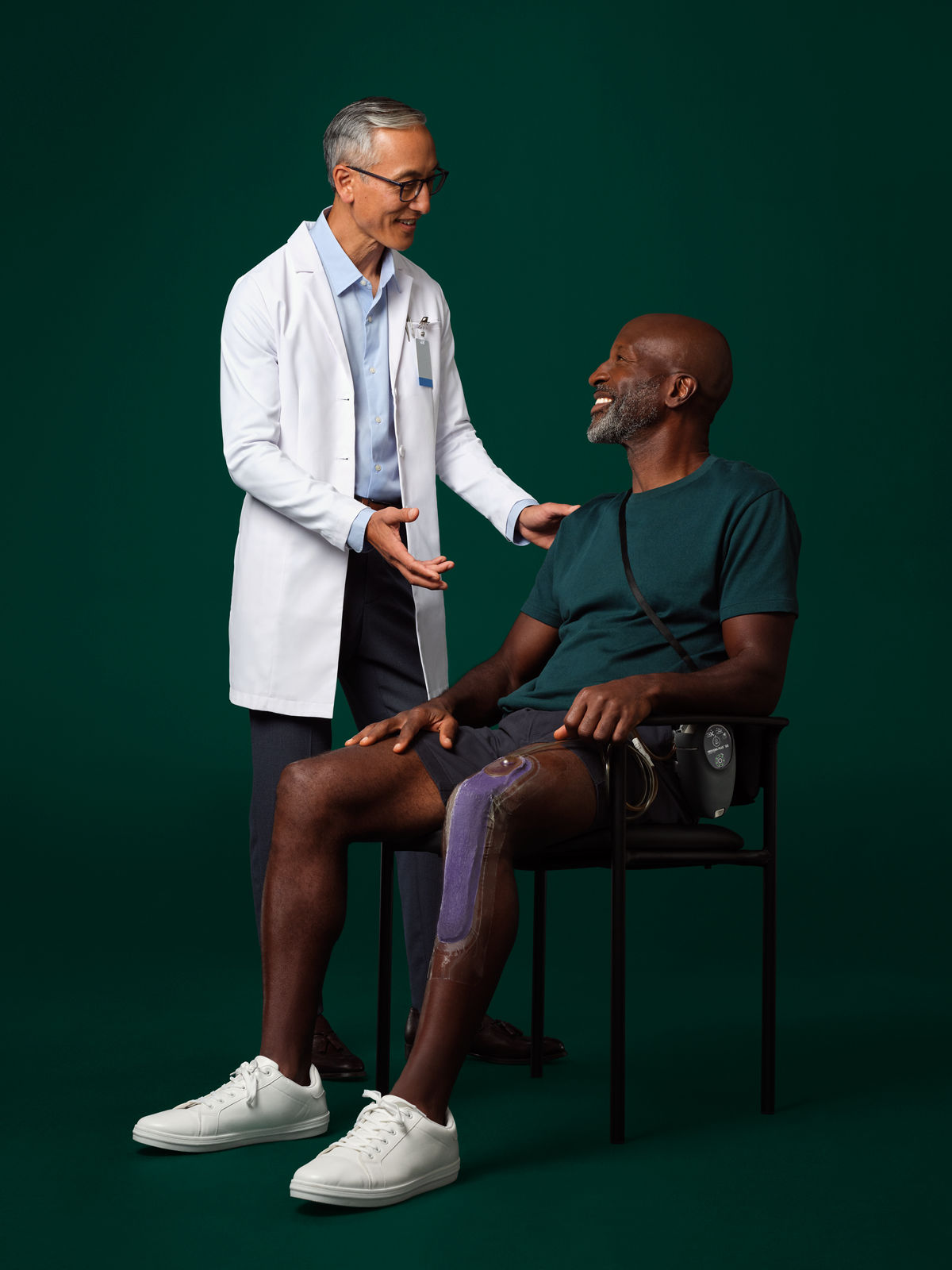A surgeon in a white coat standing next to and talking with a seated patient who has a Prevena therapy device on their leg.
