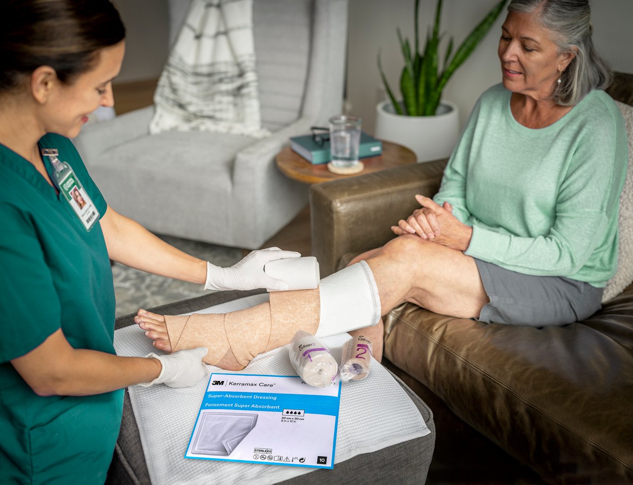 Patientin mit UCV, die mit dem zweischichtigen 3M™ Coban™ 2 Lite Kompressionssystem in der häuslichen Pflege durch einen Arzt umwickelt wird. Pakete an der Seite. 3M™ Kerramax Care™ Super-Absorbent Verband Applikation und Verpackung auch im Schuss. 