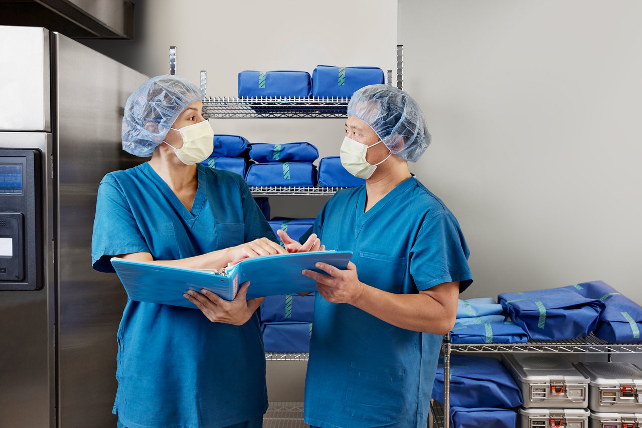 Technicians conversing and holding  binder in front of shelves next to sterilizer