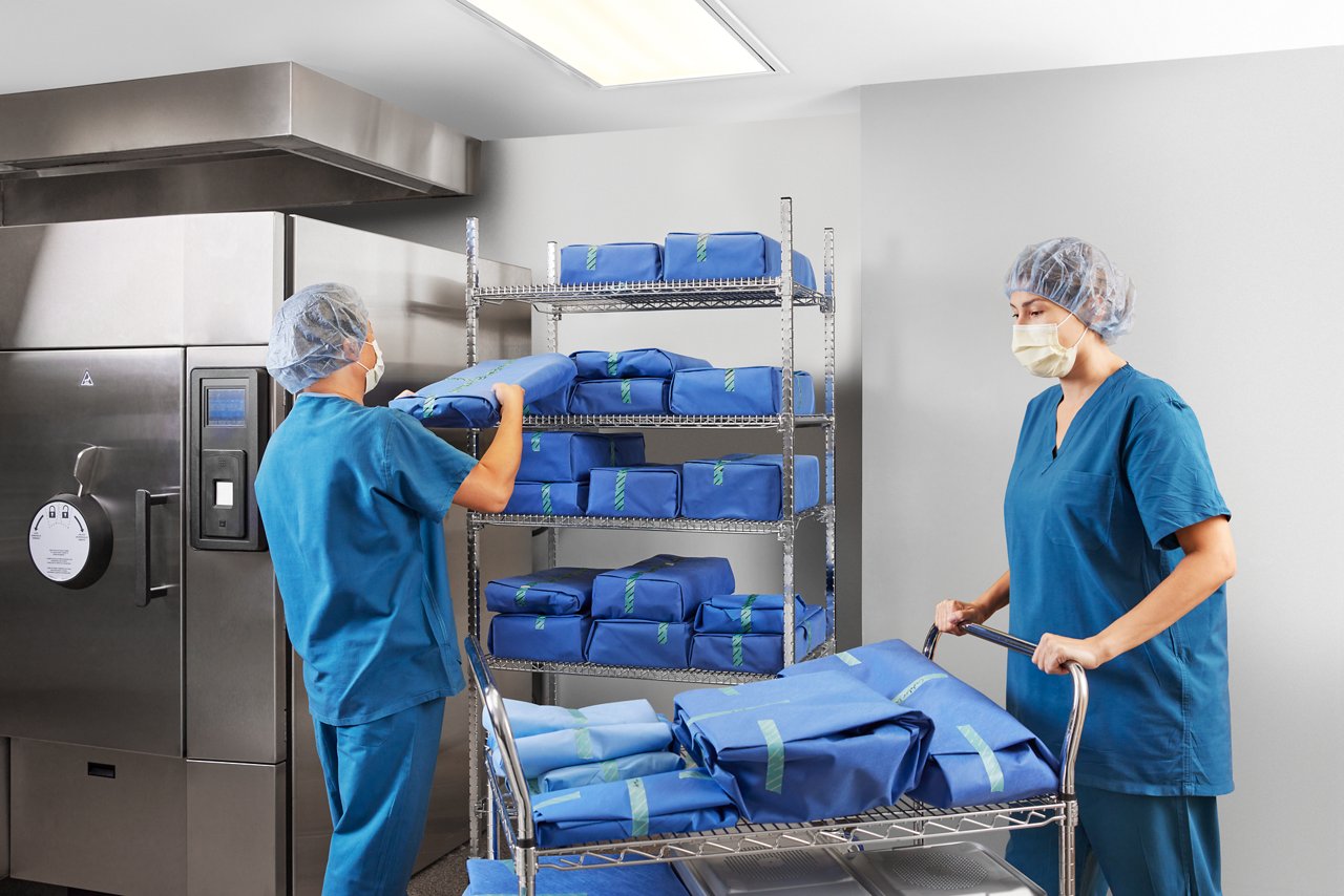 Technician holding  package in front of shelves next to sterilizer; other technician pushing trolley with packages and containers