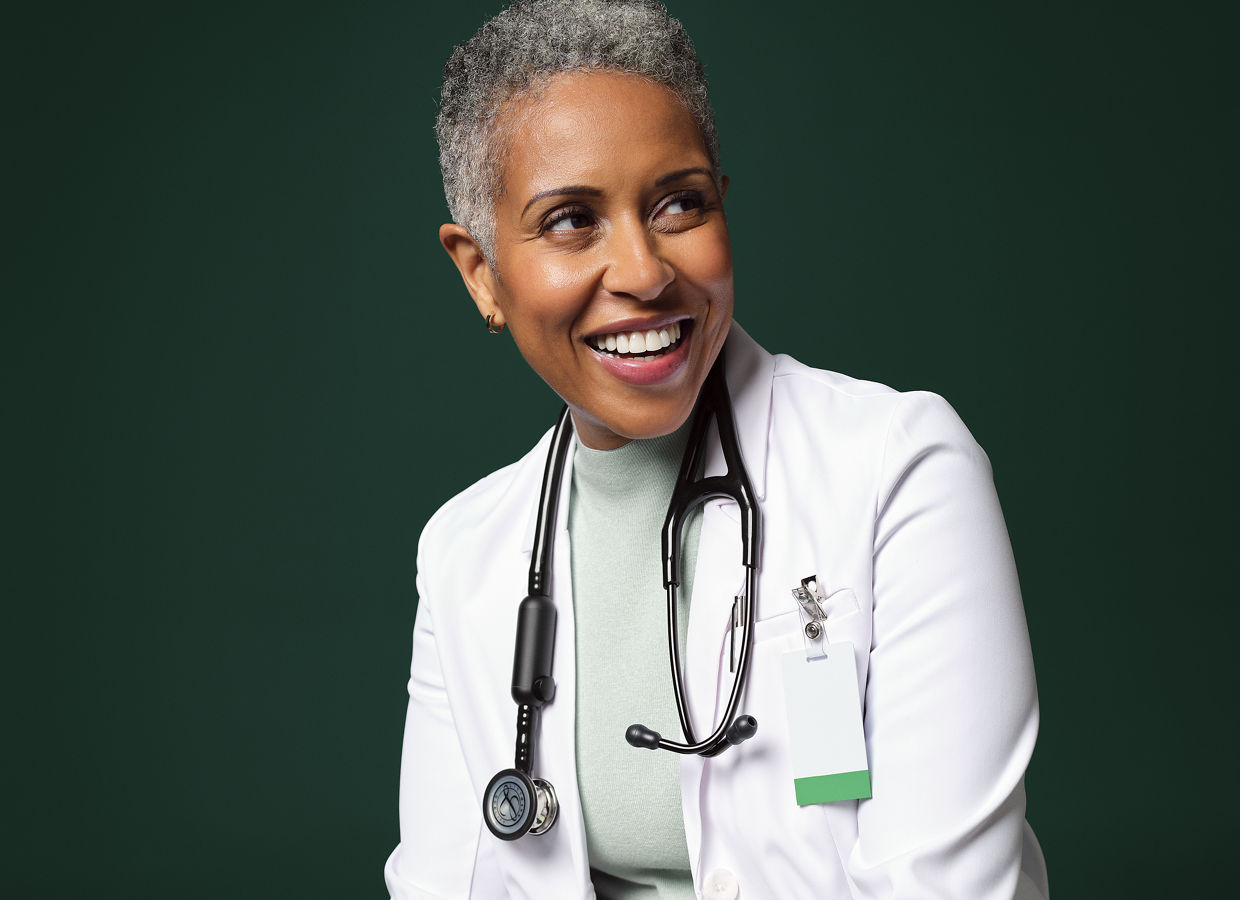 Smiling female hospital doctor seated on a stool holding a notebook.