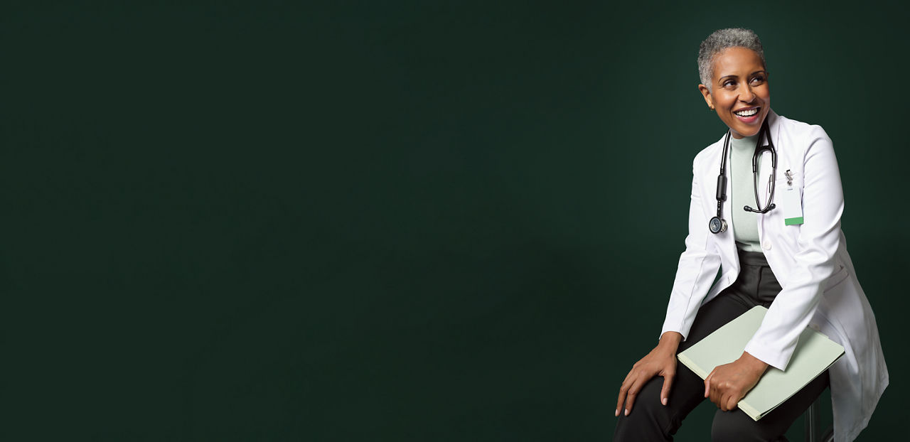 Smiling female hospital doctor seated on a stool holding a notebook.