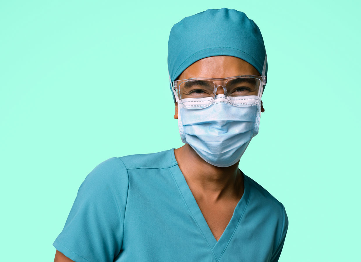Female surgical nurse in blue scrubs, cap and blue surgical mask standing with arm behind his back