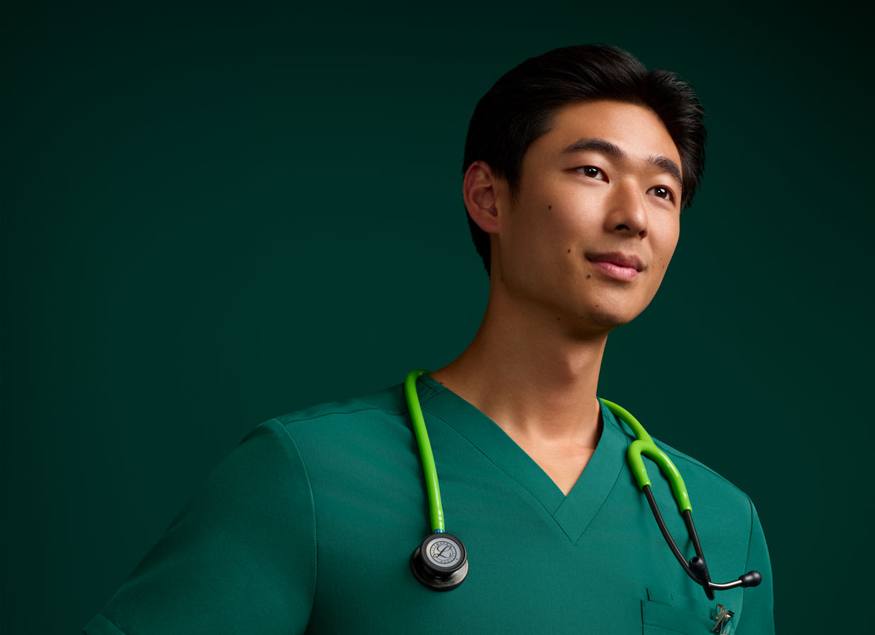 Portrait of a male nurse in scrubs standing with his hands on his hips, wearing a stethoscope against a dark green background.