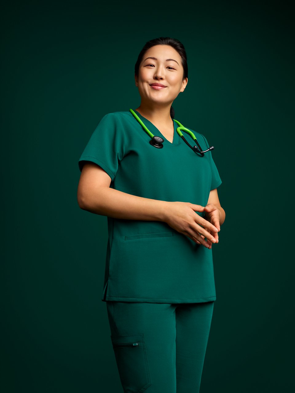 Portrait of a smiling female nurse in scrubs standing with hands together, wearing a stethoscope on a dark green background.