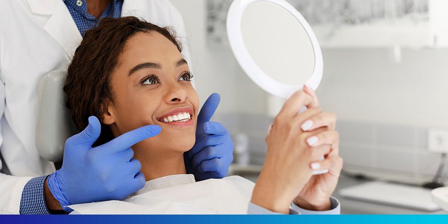 Woman in dentists chair looking in mirror and smiling with dentist pointing to her teeth, Hero Image, 900x450, rgb, jpg