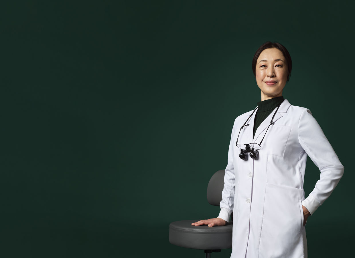 Female dentist with loupes around her neck in a white coat standing with her hand on a stool