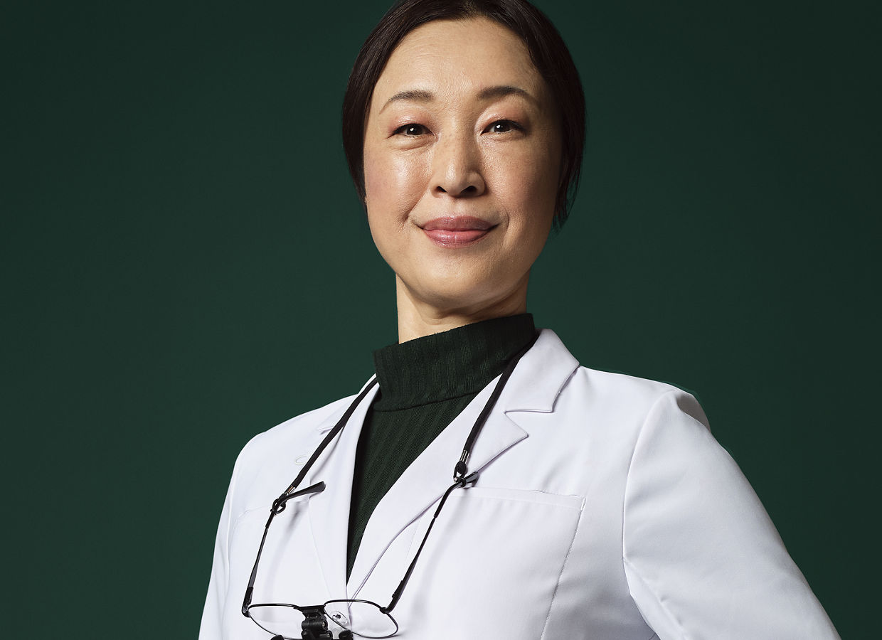 Female dentist with loupes around her neck in a white coat standing with her hand on a stool