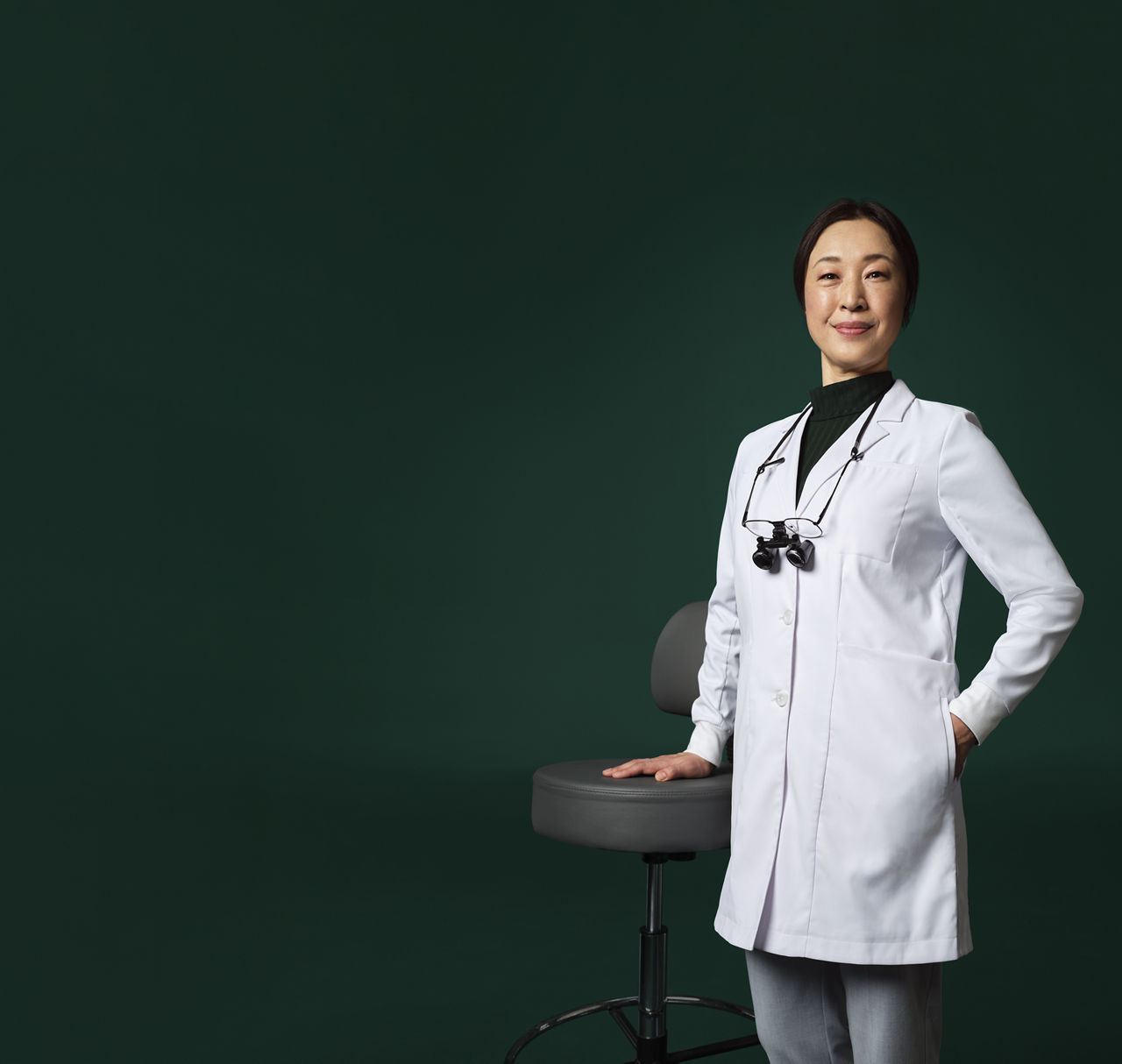 牙醫帶著放大鏡掛在脖子上，穿著白大褂，手搭在凳子上站立。