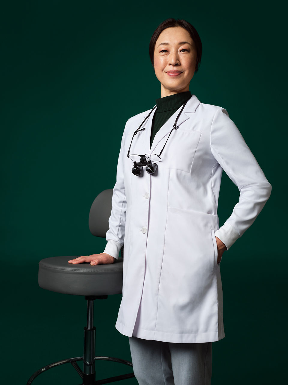 Dentist with loupes around her neck standing next to a stool.