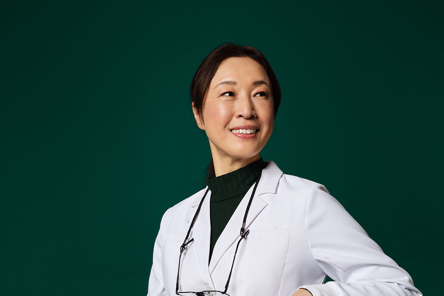 A woman in a lab coat with glasses and a loupe on a leash around her neck sits and smiles.