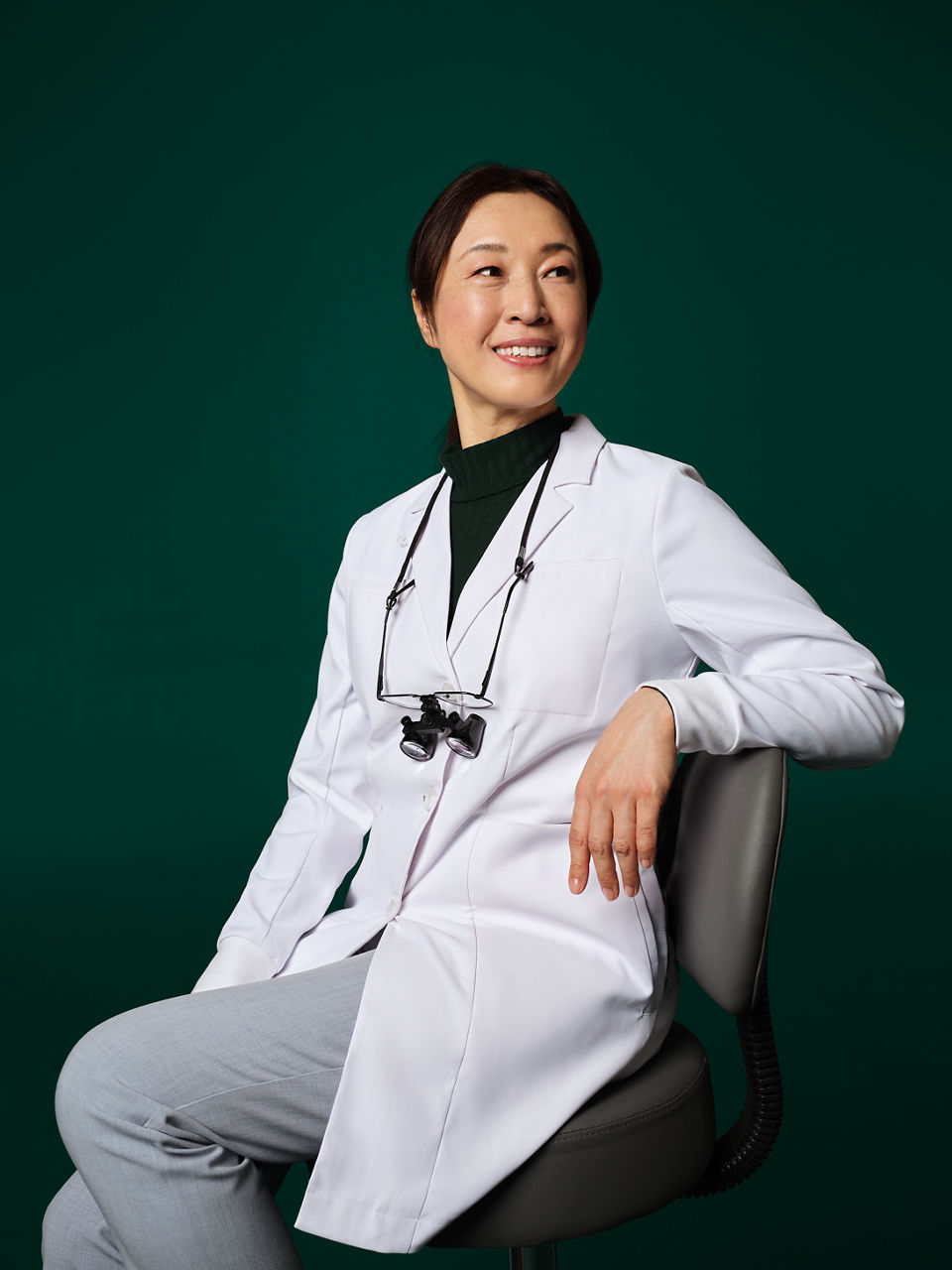 Dentist with loupes around her neck seated on a stool.