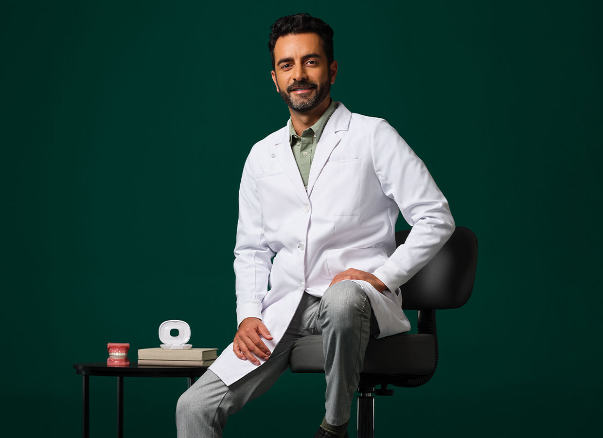 Orthodontist in a white coat sitting on a stool next to a small table displaying an aligner case and typodent.