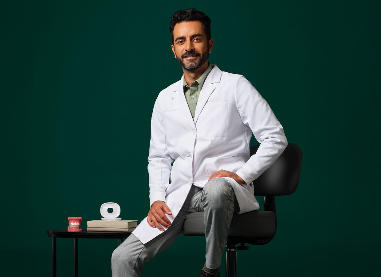 Male orthodontist wearing a white coat sitting on a stool near a small table with an aligner case and typodent on it