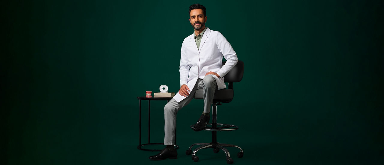 Male orthodontist wearing a white coat sitting on a stool near a small table with an aligner case and typodent on it