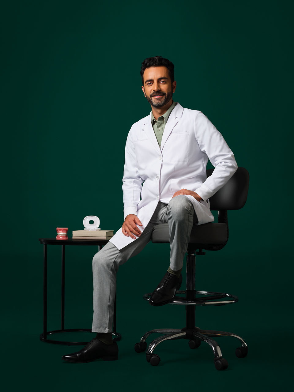 Male orthodontist wearing a white coat sitting on a stool near a small table with an aligner case and typodent on it