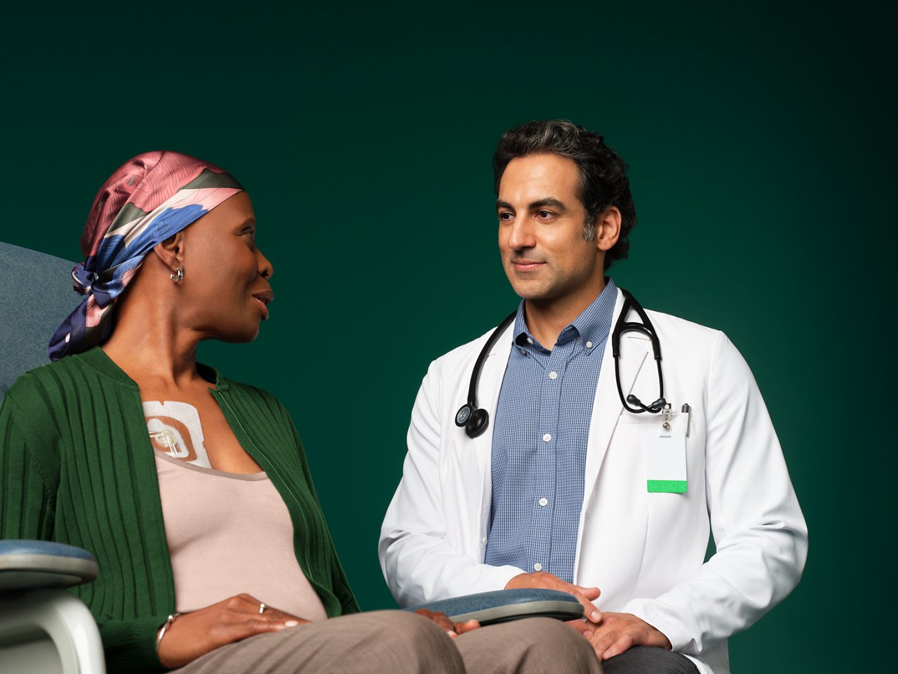 Oncologist consulting with female cancer patient sitting in hospital chair with IV port on chest covered by a Tegaderm dressing, on dark green background.