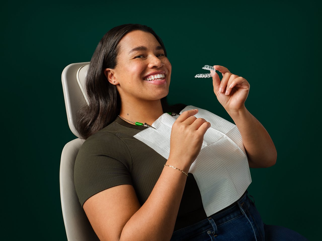 Adult female orthodontic patient seated in exam chair, smiling and holding Clarity Aligners, on a dark green background.