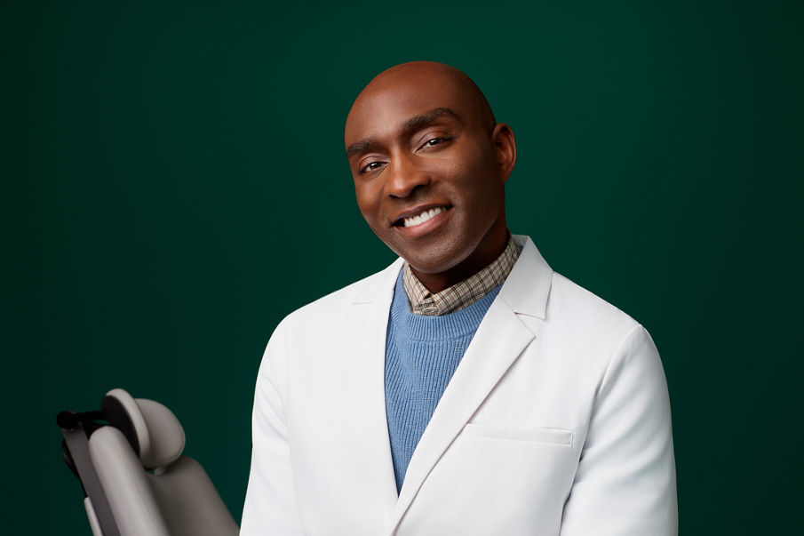 Portrait of a male orthodontist sitting next to a patient's dental chair on a dark green background.