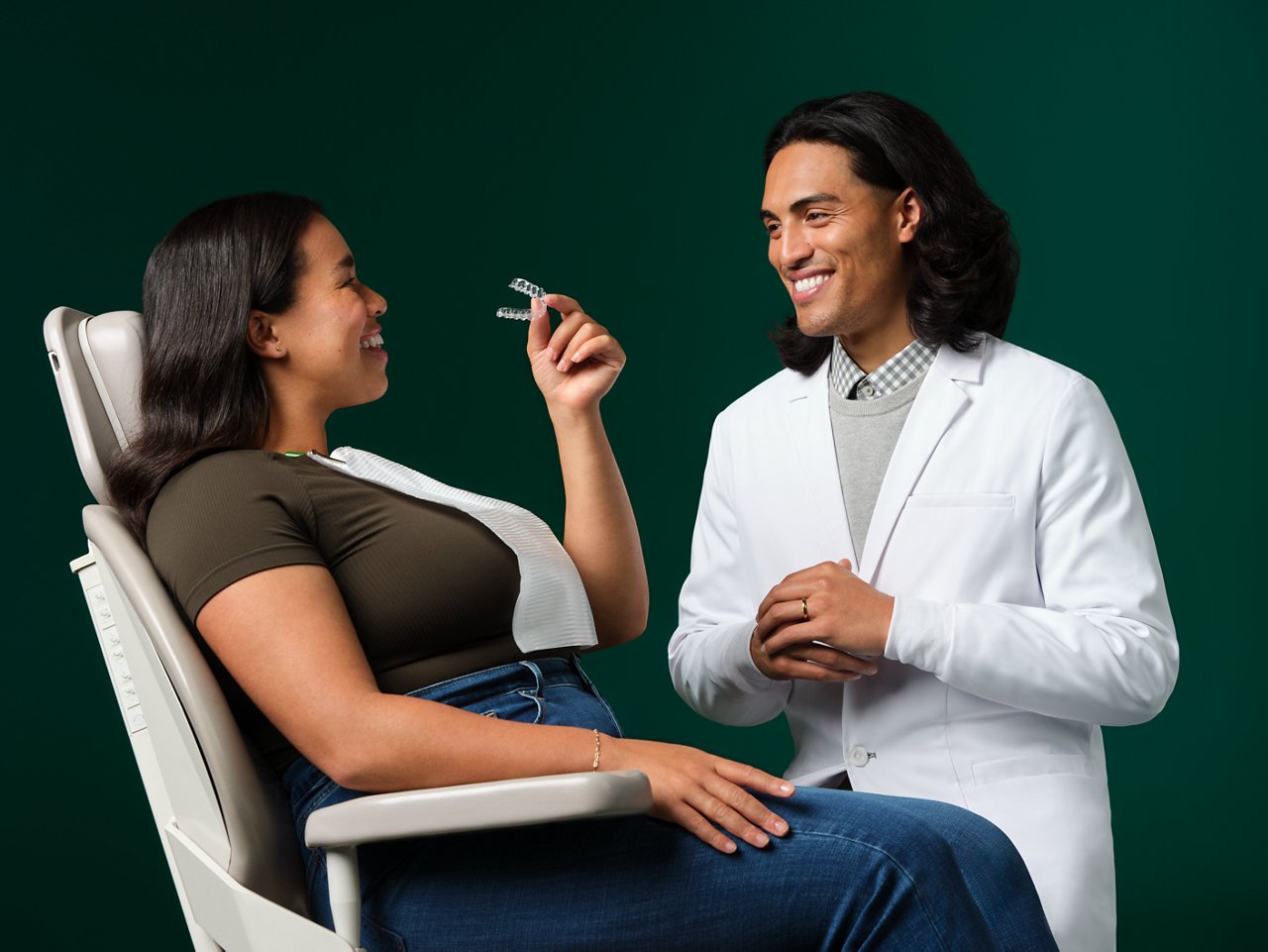 Male orthodontist consulting with an adult female orthodontic patient seated in exam chair, smiling and holding Clarity Aligners, on a dark green background.