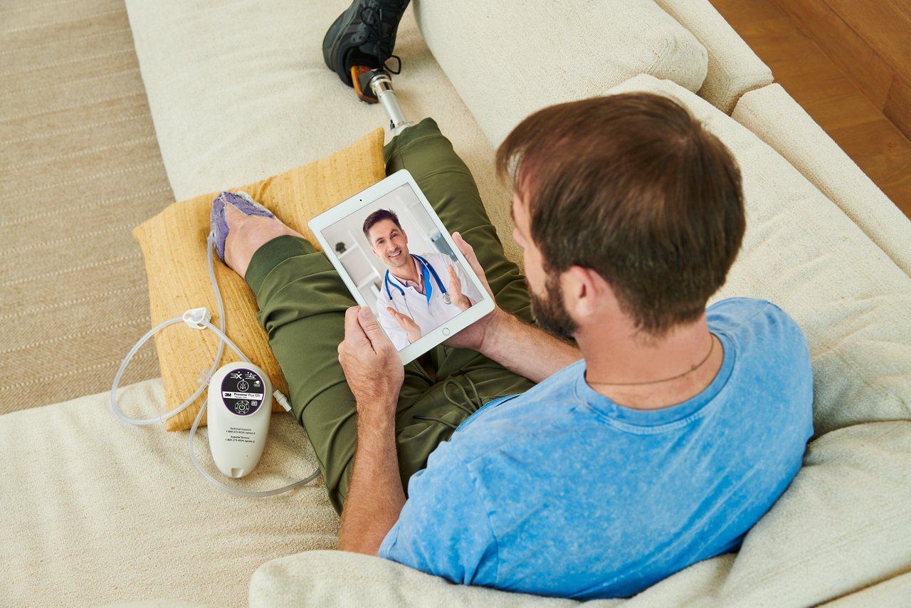 Man sitting on the couch with amputated legs, wearing the 3M™ Prevena Restor™ Adapti•Form™ dressing and the 3M™ Prevena™ Plus 125 therapy unit, talking to a telemedicine doctor on a tablet. Lifestyle image, people image, product application, high resolution, 300dpi, RGB, tiff