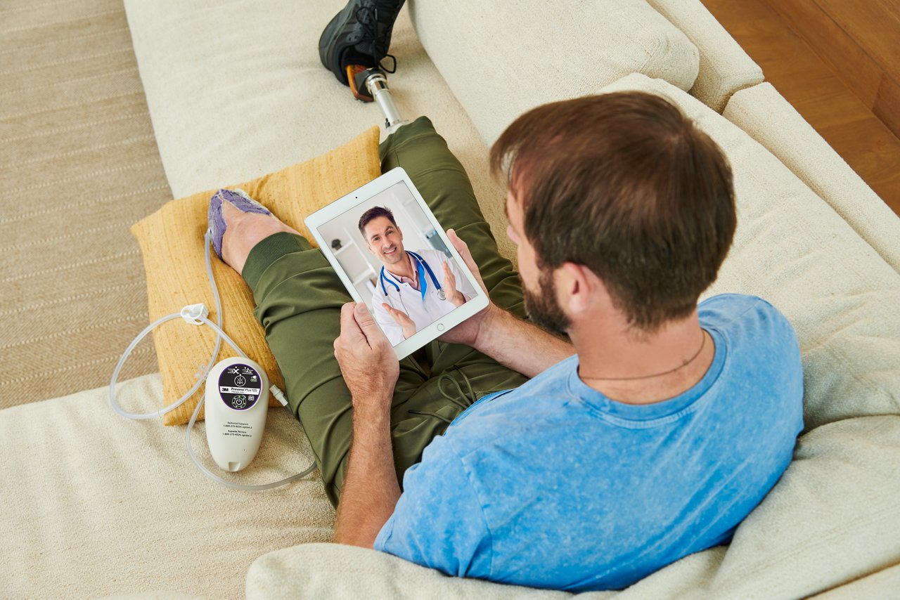 Man sitting on couch with amputated legs wearing 3M™ Prevena Restor™ Adapti•Form™ Dressing and 3M™ Prevena™ Plus 125 Therapy Unit talking with telehealth doctor on a tablet. Lifestyle image, people image, product application image, high res, 300dpi, rgb, tiff