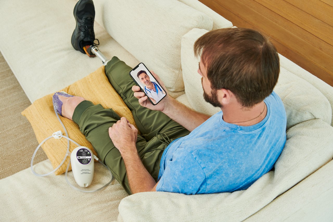 Hombre sentado en el sofá con piernas amputadas, usando el vendaje 3M™ Prevena Restor™ Adapti•Form™ y la unidad de terapia 3M™ Prevena™ Plus 125, hablando con un médico de telesalud en un smartphone. Imagen de estilo de vida, imagen de personas, imagen de aplicación de producto, alta resolución, 300dpi, rgb, tiff