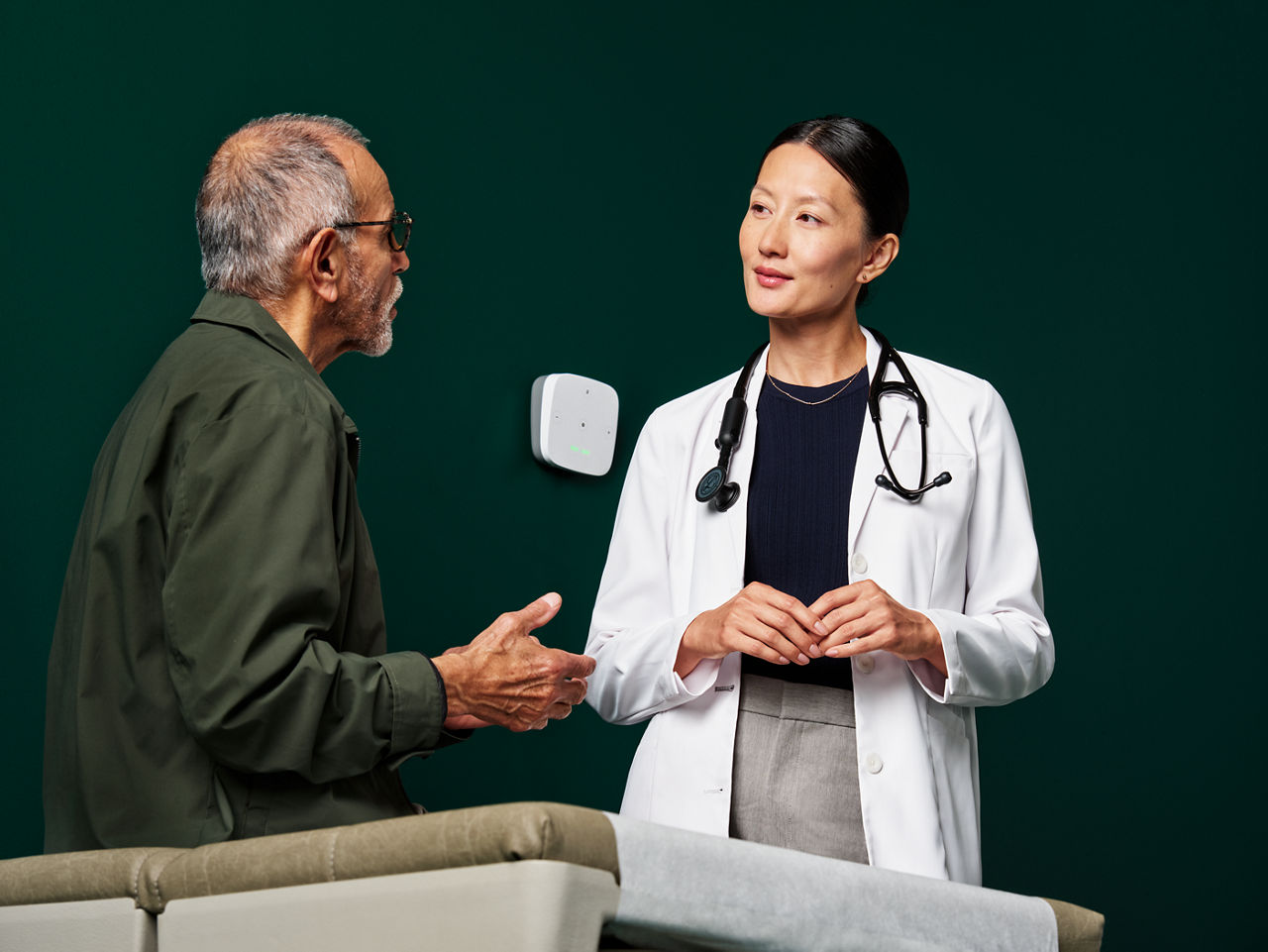 Retrato de un paciente masculino interactuando con una doctora de atención primaria en una sala de examen sobre un fondo verde oscuro.