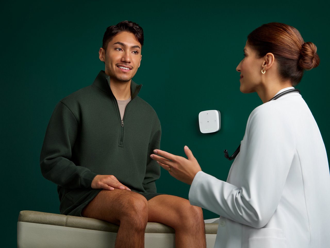 Retrato de un paciente masculino interactuando con una doctora de atención primaria en una sala de examen con un fondo verde oscuro.